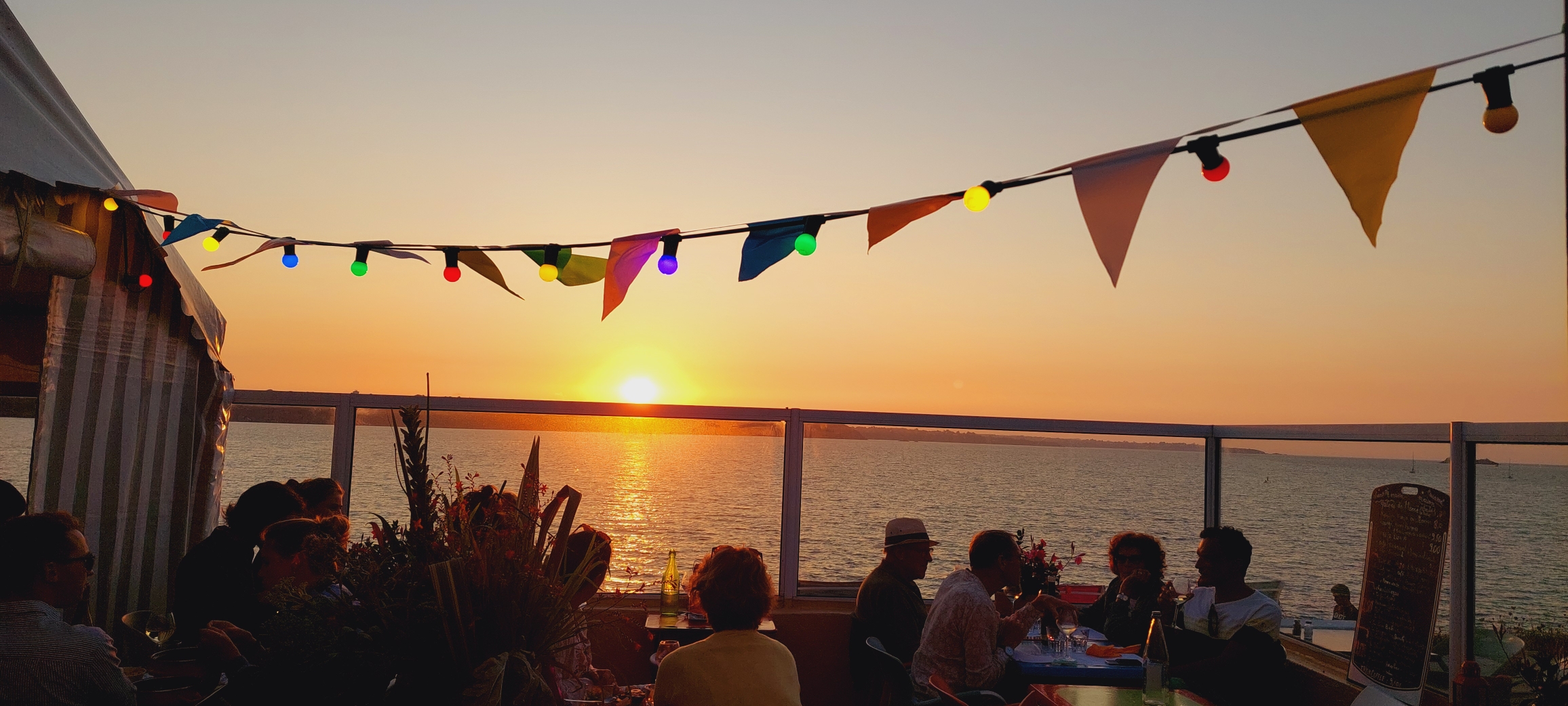 Installé face à la mer, sur la plage de la Banche à Saint-Jacut-de-la-Mer, Le Buveur de Lune est un café-restaurant chaleureux et convivial qui offre un cadre exceptionnel avec sa grande terrasse tournée vers la mer et les Ébihens. Ici, on vient avant tout profiter d’un moment de partage dans un décor breton privilégié, au rythme des marées et des couleurs du ciel. L’établissement propose une cuisine simple et généreuse autour des produits de la mer : huîtres, moules de bouchot, fruits de mer, salades, tapas et planches à partager. À l’heure de l’apéritif, la terrasse devient un véritable lieu de détente, particulièrement apprécié pour les cocktails au coucher du soleil, avec une vue dégagée sur la baie et les parcs à huîtres et à moules. Avec son ambiance estivale, sa musique, son esprit décontracté et son panorama remarquable, le Buveur de Lune est une adresse idéale pour déjeuner, dîner, boire un verre ou simplement profiter d’un coucher de soleil face à la mer. Le Buveur de Lune, c’est avant tout une expérience : la mer, les produits locaux, les amis et le plaisir de prendre le temps.